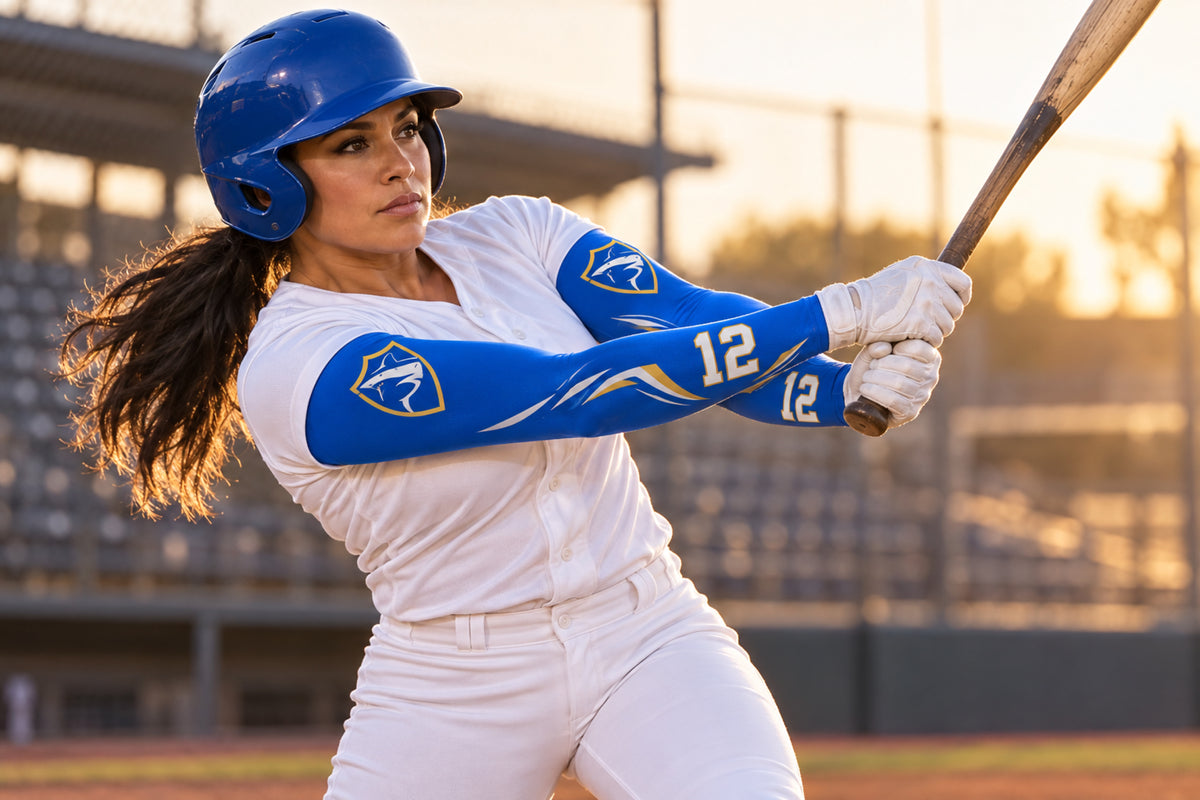 Batter finishing a two-hand swing in Royal Blue custom arm sleeves with a shark crest and number 12